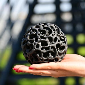 Puede incluir: Una esfera porosa de color gris oscuro con un diseño intrincado y abierto, sostenida en una mano. La superficie de la esfera tiene numerosos agujeros. El fondo está borroso, con toques de verde y una estructura metálica.