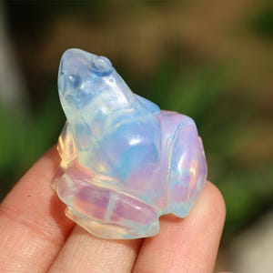 May include: A small, iridescent opal frog figurine. The frog is carved from a translucent, milky white opal with blue and purple iridescence.