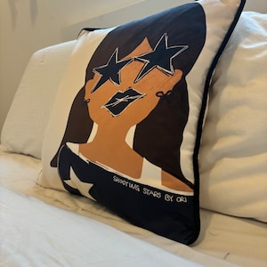 Könnte beinhalten: Ein weißes Kissen mit einer schwarzen und braunen Illustration einer Person, die eine sternförmige Sonnenbrille trägt. Der Text "Shooting Stars by ORJ" ist auf dem Kissen aufgedruckt.