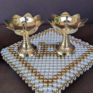 May include: Two gold-colored, lotus-shaped oil lamps sit atop a square, beaded decorative mat. The mat is adorned with white and gold-colored beads. The lamps have a pedestal base and a small, central holder for the wick.