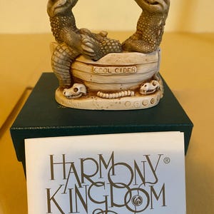 May include: A detailed figurine featuring two alligator-like creatures in a wooden barrel with the text "COOL CIDER". The figurine sits on a green box, with a card that reads "HARMONY KINGDOM" and "The Original Box Figurine".