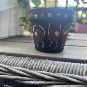 Puede incluir: Una maceta de terracota negra con un borde blanco y rojo. Tres caras de dibujos animados marrones están pintadas en la parte delantera de la maceta.