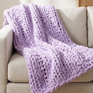 May include: A chunky knit throw blanket in a solid light lavender color. The blanket features a thick, hand-knitted texture with large, interlocking loops of soft yarn, draped over the arm and seat of a neutral-toned upholstered sofa.