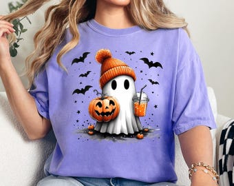 Op de afbeelding: Een lichtpaars T-shirt met een cartoongeest met een oranje muts, met een uitgesneden pompoen en een drankje. Zwarte vleermuizen en sterren omringen de geest en creëren een Halloween-thema.