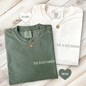 May include: Two t-shirts with the Roman numeral date XII.XXIV.MMIII printed on them. One shirt is ivory and the other is moss green.