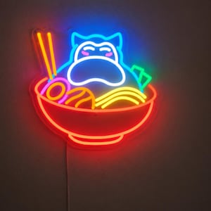 Peut inclure: Une enseigne néon représentant un personnage de dessin animé aux yeux fermés et aux joues roses, assis dans un bol de ramen rouge. Le design inclut des baguettes jaunes, des nouilles jaunes, une spirale violette et des algues vertes.