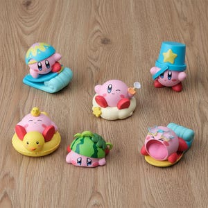 May include: Six pink Kirby figurines in summer poses. One wears a blue helmet and rides a surfboard, another holds a sand shovel, and others are in a duck floatie, watermelon hat, and sunglasses. Figures are on a wooden surface.