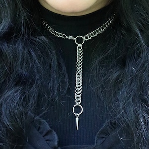 May include: A close-up of a person wearing a black choker necklace with a silver chain and a pointed pendant. The necklace is made of a thick chain with a ring at the top and a long, thin chain with a pointed pendant hanging from it. The person is wearing a black shirt with a lace-up detail.