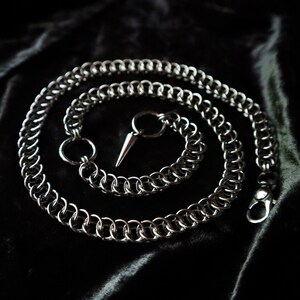 May include: A silver chain with a pointed tip and a clasp. The chain is coiled on a black fabric background.