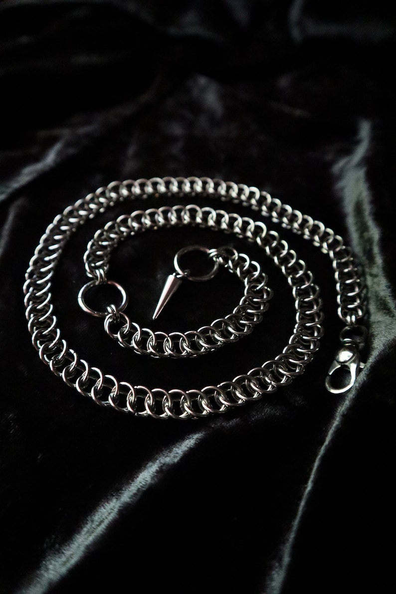 May include: A silver chain with a pointed tip and a clasp. The chain is coiled on a black fabric background.