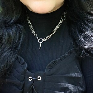 May include: A close-up of a person wearing a black mock neck shirt with a silver chain necklace featuring a pointed pendant. The shirt has a ruffled neckline and a lace-up front with silver eyelets.