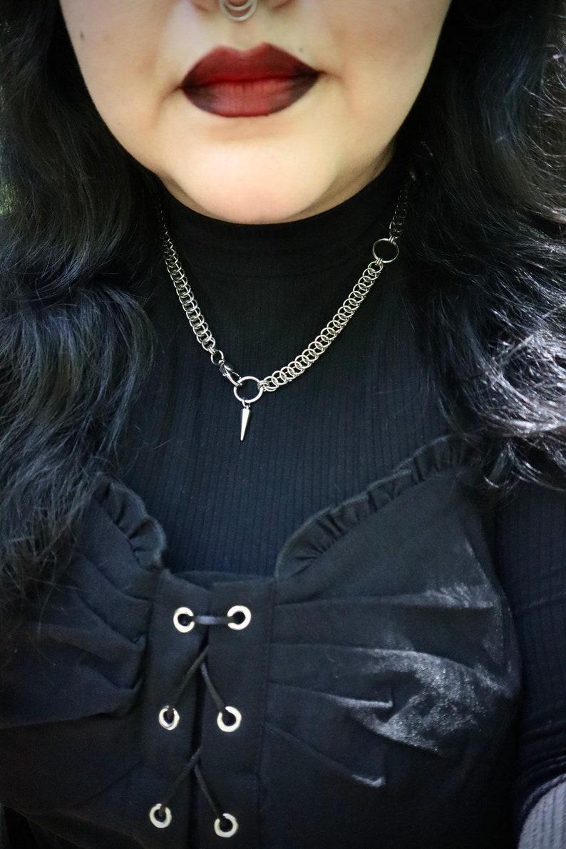 May include: A close-up of a person wearing a black mock neck shirt with a silver chain necklace featuring a pointed pendant. The shirt has a ruffled neckline and a lace-up front with silver eyelets.