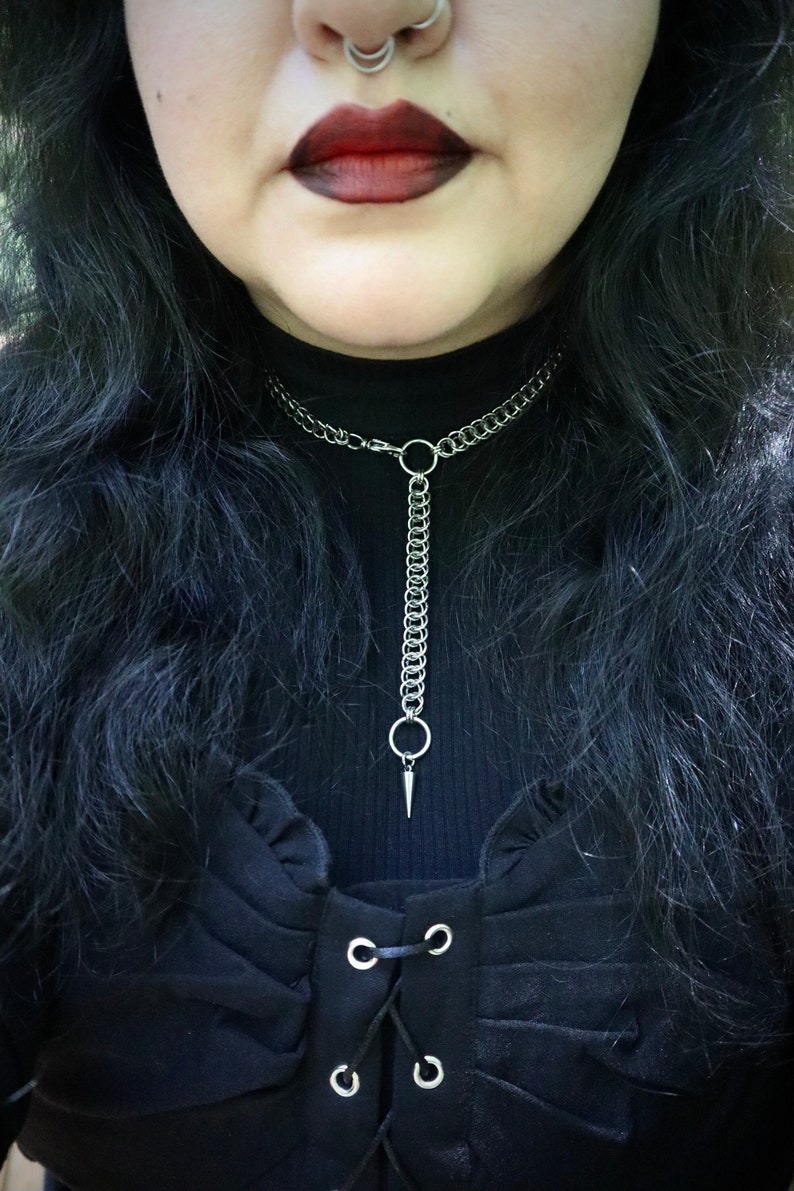 May include: A close-up of a person wearing a black choker necklace with a silver chain and a pointed pendant. The necklace is made of a thick chain with a ring at the top and a long, thin chain with a pointed pendant hanging from it. The person is wearing a black shirt with a lace-up detail.