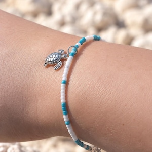 May include: A beaded bracelet featuring alternating white and turquoise seed beads. The bracelet includes a silver-toned metal charm shaped like a sea turtle, attached to the strand with a small circular jump ring, and a metal chain extender.