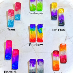 Pode incluir: Um conjunto de 10 pares de brincos retangulares coloridos, cada par representando uma bandeira de orientação sexual diferente. As bandeiras incluem: transgênero, genderqueer, não binário, arco-íris, bissexual, assexual, lésbica.