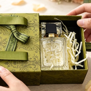 May include: A rectangular olive green gift box with a decorative paisley pattern and matching ribbon handles. Inside, a clear glass perfume bottle with a black cap rests on shredded paper filler. The bottle features white cursive text on the front.