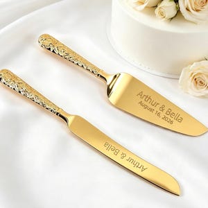Puede incluir: Un juego de cuchillo y servidor de pastel dorado. El servidor tiene una hoja ancha y plana, y el cuchillo tiene una hoja larga y estrecha. Ambos tienen mangos ornamentados con diseños florales. El servidor está grabado con "Arthur & Bella August 16, 2026".