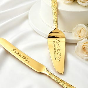 Puede incluir: Juego de cuchillo y servidor de pastel dorados con detalles florales en el mango. El servidor y el cuchillo están grabados con los nombres "Noah & Chloe" y la fecha "20 de marzo de 2026". El juego se muestra sobre una superficie blanca, cerca de un pastel blanco y rosas.