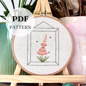 May include: A PDF pattern for embroidery. The design features a pink flower with green leaves and two bees, framed within a black line drawing. The embroidery is set in a wooden hoop, displayed on a small wooden easel. The text "PDF PATTERN" is visible.