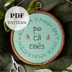 May include: A round embroidery hoop with a light teal fabric featuring the text "Mash Em, Stick Em, Boil Em, Put Em In A Stew, Po Ta Toes" in brown thread. The hoop is made of wood and has a metal clasp. The image also includes a pair of scissors.