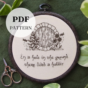 May include: A framed embroidery hoop featuring a hand-stitched design of a hobbit door with the text "In a hole in the ground there lived a hobbit." The text "PDF PATTERN" is overlaid on the image. The frame is dark brown.