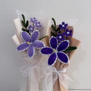 Op de afbeelding: Twee boeketten van handgemaakte, paarse bloemstukken. Elk boeket heeft grote, vijfbladige bloemen met witte randen, kleinere paarse bloemen en groene bladeren. De boeketten zijn verpakt in bruin papier en vastgebonden met witte linten.