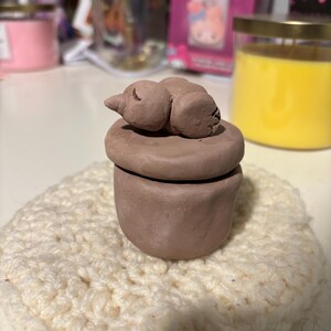 May include: A handmade, brown clay trinket box with a sleeping animal figurine on top. The box is sitting on a cream-colored, crocheted coaster. The background includes candles and other decorative items.