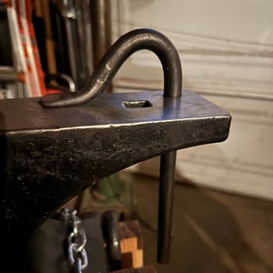 May include: A black metal anvil with a metal hook attached to the top. The hook is bent into a U shape and is attached to the anvil with a metal rod. The anvil is sitting on a wooden base with a chain attached to the side.