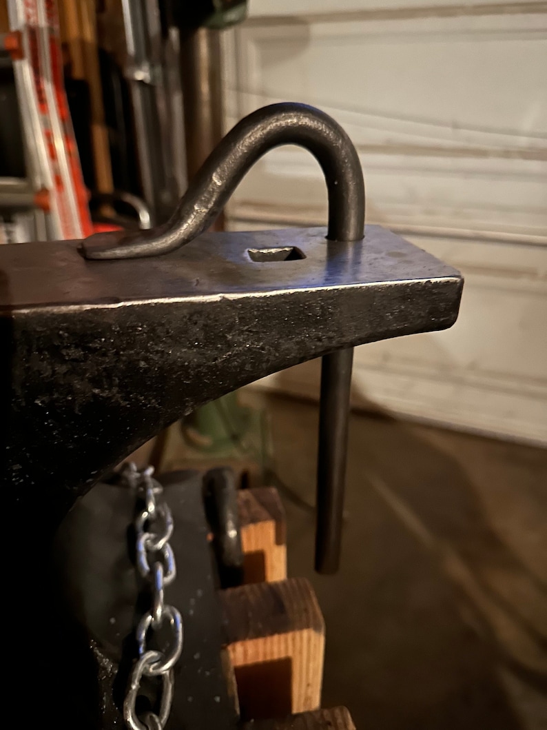 May include: A black metal anvil with a metal hook attached to the top. The hook is bent into a U shape and is attached to the anvil with a metal rod. The anvil is sitting on a wooden base with a chain attached to the side.