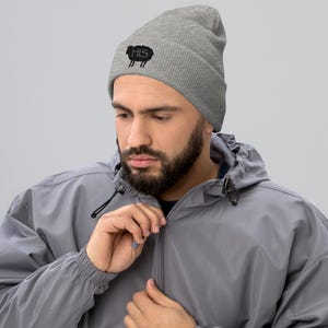 Pode incluir: Um gorro cinza com a palavra "HIS" e um gráfico de ovelha preta. O gorro é feito de um material de malha canelada macia. A pessoa está usando uma jaqueta cinza.