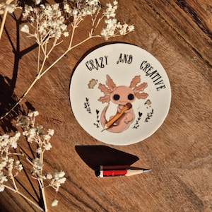 Peut inclure: Une petite assiette ronde en céramique avec les mots "CRAZY AND CREATIVE" encerclant un axolotl de dessin animé tenant un crayon. Un crayon miniature avec des rayures rouges et noires est à proximité. Le fond est une surface en bois avec des fleurs blanches.