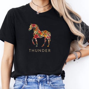 Op de afbeelding: Zwart T-shirt met een bloemenpaardenontwerp en het woord "THUNDER" eronder.