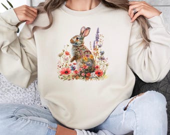 Può includere: Una felpa color crema con un'illustrazione ad acquerello di un coniglio marrone circondato da fiori colorati. Il coniglio è seduto in un campo di fiori selvatici.