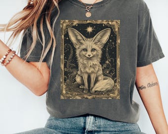 May include: Dark gray t-shirt featuring a detailed illustration of a fennec fox within a decorative frame. The artwork is in sepia tones, set against a dark background with celestial elements. The shirt is a casual wear item.