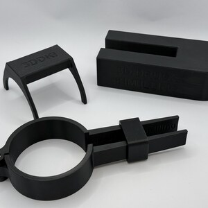 Könnte beinhalten: Schwarze 3D-gedruckte Objekte, darunter ein kleiner Tisch mit dem Text "3DDOKI", ein U-förmiger Block mit Text und eine kreisförmige Klemme mit einem verstellbaren Arm. Die Objekte befinden sich auf einer weißen Oberfläche.