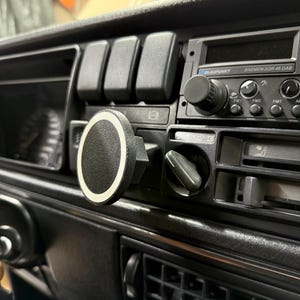 Könnte beinhalten: Nahaufnahme eines schwarzen Armaturenbretts mit Radio und Lüftungsschlitzen. Eine runde, schwarze Telefonhalterung mit weißem Rand ist angebracht. Das Radio hat ein digitales Display und mehrere Knöpfe. Das Armaturenbrett besteht aus schwarzem Kunststoff.