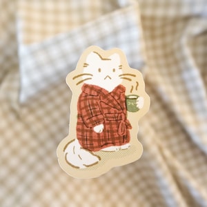 May include: A white cat wearing a red and black plaid robe holds a green mug with steam coming from it.