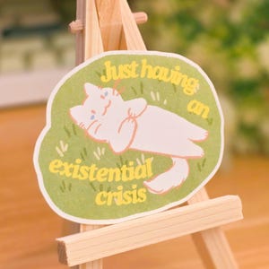 May include: A green and yellow sticker with a white cat lying on its back with the text "Just having an existential crisis". The sticker is on a small wooden easel.