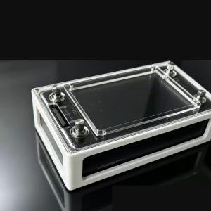 Può includere: Un dispositivo elettronico rettangolare con una cornice esterna bianca e un pannello di controllo nero. La superficie superiore include una copertura in acrilico trasparente, uno schermo centrale e quattro manopole metalliche.