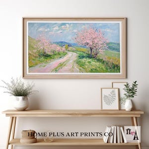 K&ouml;nnte beinhalten: Ein gerahmtes Gem&auml;lde, das eine Landstra&szlig;e zeigt, die sich durch ein Feld mit bl&uuml;henden rosa B&auml;umen schl&auml;ngelt. Der Himmel ist blau mit wei&szlig;en Wolken. Das Gem&auml;lde ist in einem hellen Holzrahmen.