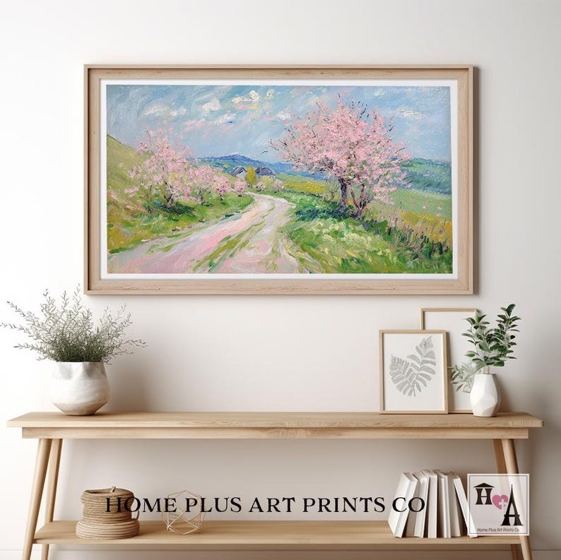 K&ouml;nnte beinhalten: Ein gerahmtes Gem&auml;lde, das eine Landstra&szlig;e zeigt, die sich durch ein Feld mit bl&uuml;henden rosa B&auml;umen schl&auml;ngelt. Der Himmel ist blau mit wei&szlig;en Wolken. Das Gem&auml;lde ist in einem hellen Holzrahmen.