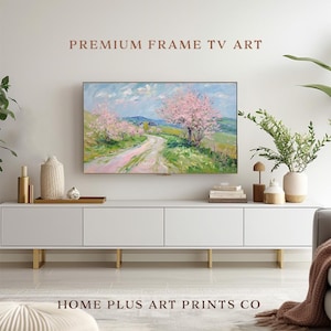 K&ouml;nnte beinhalten: Eine wei&szlig;e Entertainment-Konsole mit goldenen Beinen, die einen gerahmten Leinwanddruck eines Landschaftsgem&auml;ldes mit rosa bl&uuml;henden B&auml;umen und einem gewundenen Weg zeigt. Der Text "PREMIUM FRAME TV ART" wird &uuml;ber dem Gem&auml;lde angezeigt und "HOME PLUS ART PRINTS CO" unter der Konsole.