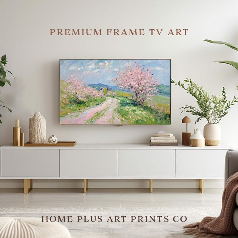 K&ouml;nnte beinhalten: Eine wei&szlig;e Entertainment-Konsole mit goldenen Beinen, die einen gerahmten Leinwanddruck eines Landschaftsgem&auml;ldes mit rosa bl&uuml;henden B&auml;umen und einem gewundenen Weg zeigt. Der Text "PREMIUM FRAME TV ART" wird &uuml;ber dem Gem&auml;lde angezeigt und "HOME PLUS ART PRINTS CO" unter der Konsole.