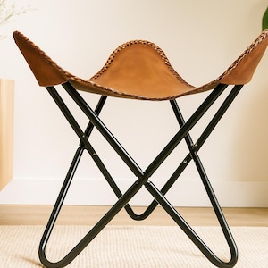 May include: A brown leather butterfly chair with a black metal frame. The chair has a curved seat and a stitched edge. The legs are angled and meet in the center. The chair is a stylish piece of furniture.