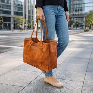 Peut inclure: Un grand sac fourre-tout rectangulaire en cuir marron avec deux poignées et une poche extérieure. Le sac est porté par une personne portant un jean bleu et des chaussures plates beiges sur un trottoir.