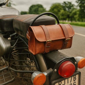 May include: A brown leather motorcycle saddlebag with two straps and brass buckles is mounted on the back of a motorcycle. The motorcycle is silver and black, with a black seat and a red taillight. The background is a blurred road and trees.