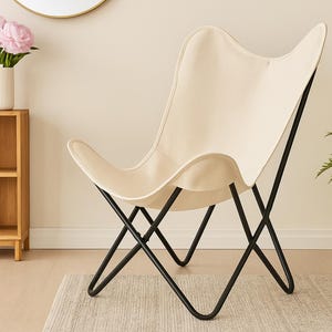 Puede incluir: Silla mariposa crema con estructura de metal negro. La silla se encuentra contra una pared de tonos neutros, con un mueble de madera, un jarrón con flores rosas y una planta en maceta en el fondo. El diseño de la silla es moderno y minimalista.