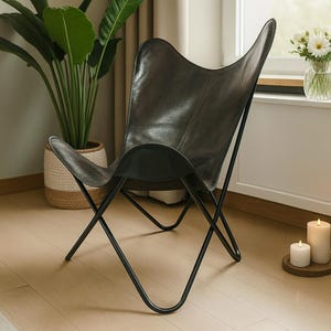 Puede incluir: Una silla mariposa de cuero negro con estructura de metal negro. La silla está en una habitación con una planta en maceta grande, velas y un jarrón con flores blancas. Sus dimensiones son de aproximadamente 76 cm de ancho, 89 cm de alto y 76 cm de profundidad.