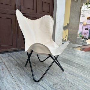 Puede incluir: Una silla mariposa crema con estructura de metal negro. La silla tiene un diseño distintivo en forma de ala. Se encuentra frente a una puerta marrón y una superficie embaldosada. Esta moderna silla es adecuada para uso interior o exterior.