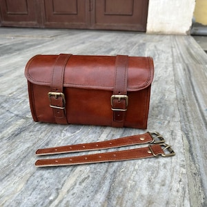 Może przedstawiać: Brązowa skórzana torba z dwoma paskami i mosiężnymi klamrami. Torba jest prostokątna z zakrzywionym wierzchem, przeznaczona do roweru lub motocykla. Dołączone są dwa dodatkowe skórzane paski z klamrami.